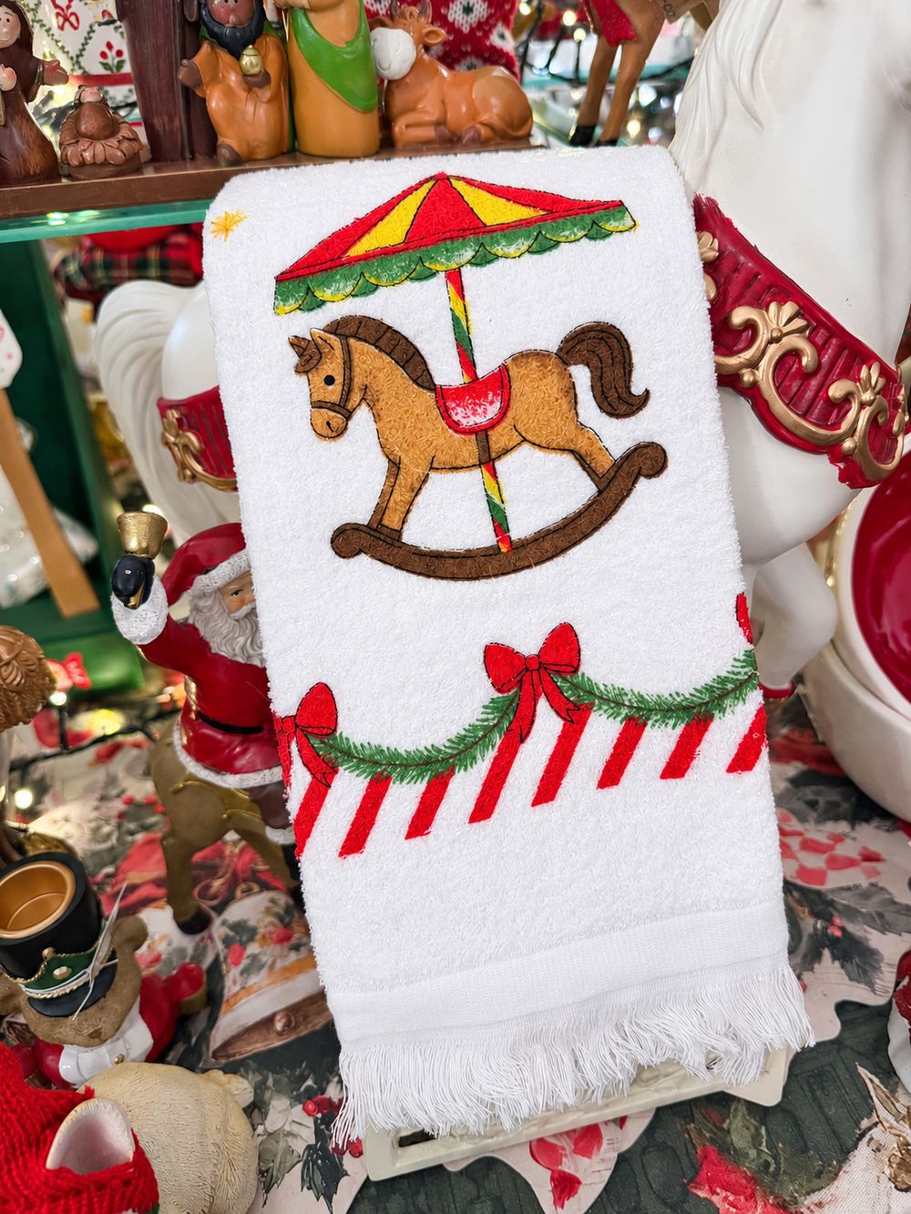 Essuie-main Carrousel de Noël blanc à franges, présenté plié dans le décor de la boutique, vue 1.