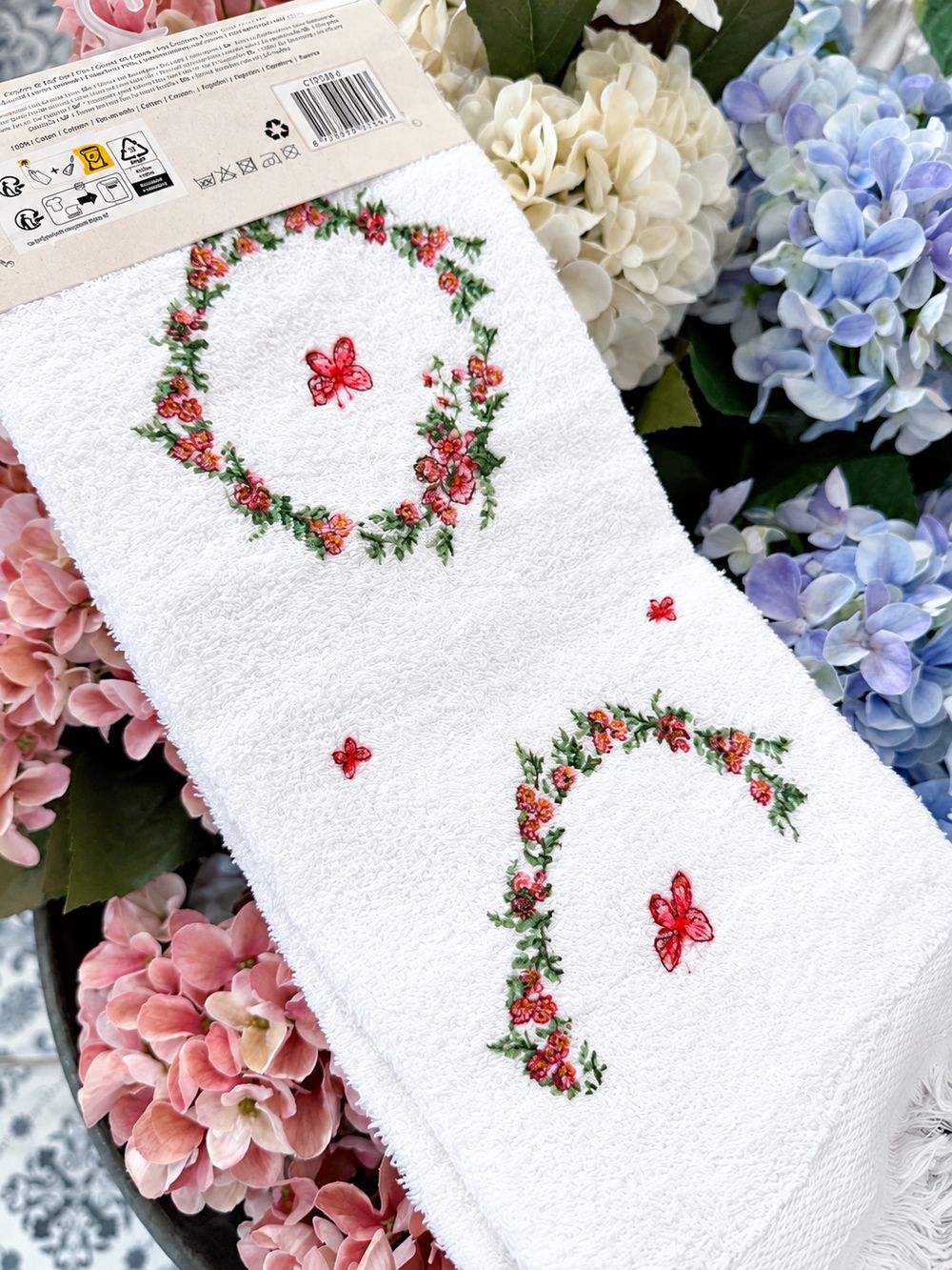 Détail des couronnes florales et papillons sur l’essuie-mains blanc en coton, parmi des hortensias.