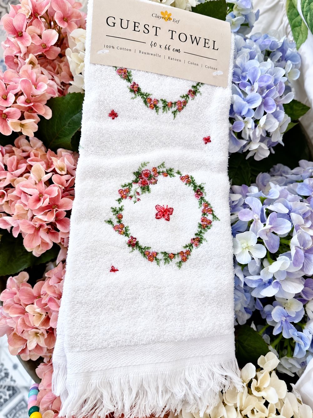 Vue de face de l’essuie-mains Couronne Florale avec son emballage et ses franges, entouré d’hortensias.