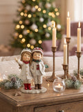 Figurine de Noël représentant deux enfants enlacés en tenue d’hiver, dans des tons blanc cassé, rouge et vert, idéale pour une décoration de Noël romantique et poétique