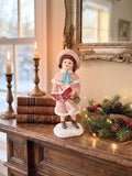 Figurine de Noël représentant une petite chanteuse en manteau rose et bonnet assorti, dans un style victorien romantique, sur fond de décoration shabby chic