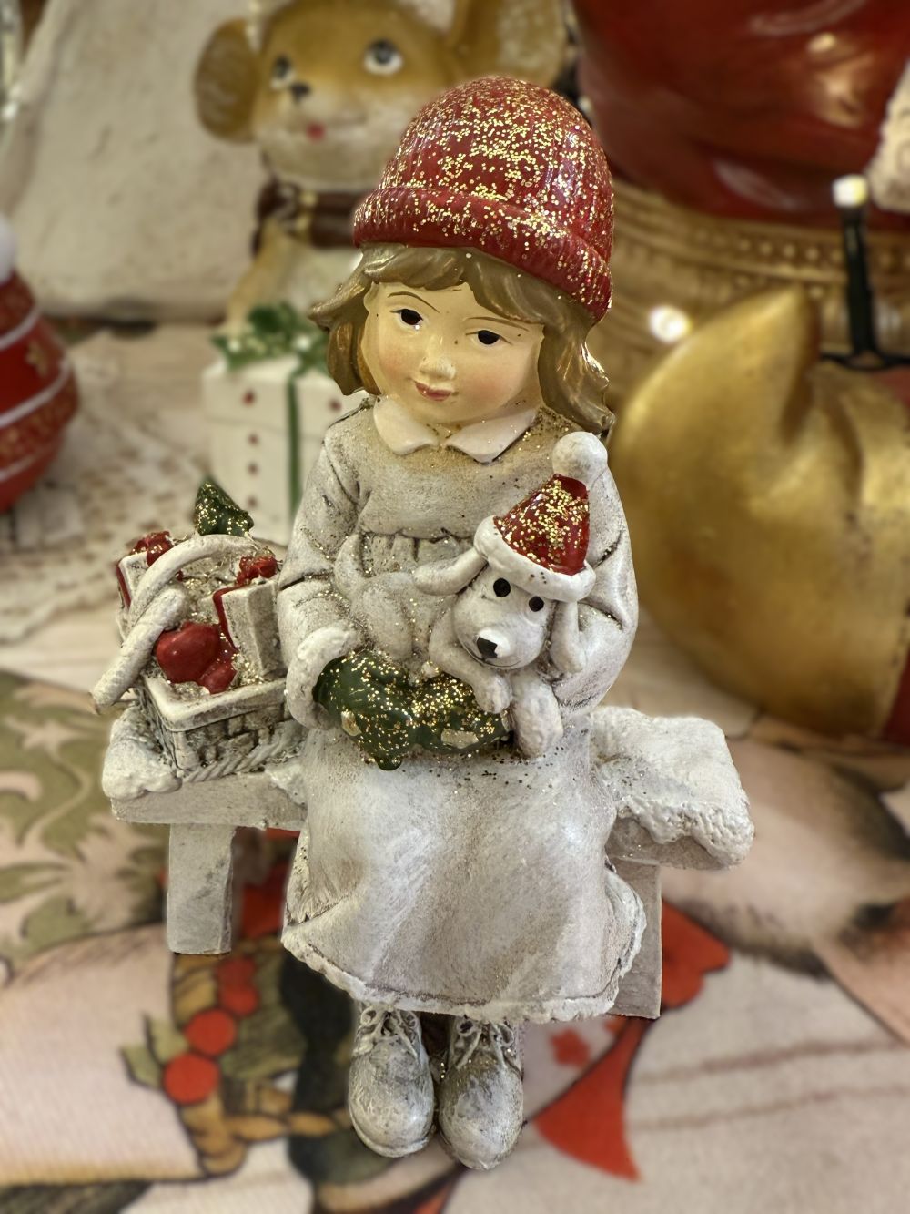 Figurine de Noël représentant une fillette assise sur un banc avec son petit chien blanc et un panier de cadeaux, couleurs blanc rouge et beige, style décoration romantique shabby chic et ambiance féérique