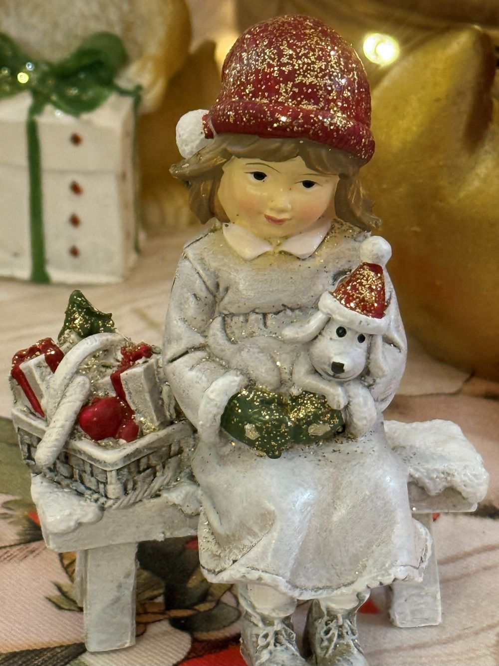 Figurine de Noël représentant une fillette assise sur un banc avec son petit chien blanc et un panier de cadeaux, couleurs blanc rouge et beige, style décoration romantique shabby chic et ambiance féérique