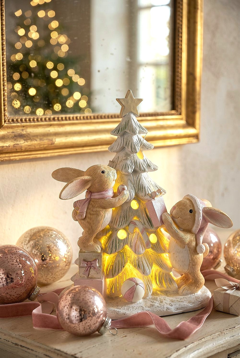 Figurine de Noël avec deux lapinous autour d’un sapin lumineux, tons vert givré, crème et rose poudré, décoration féérique et romantique