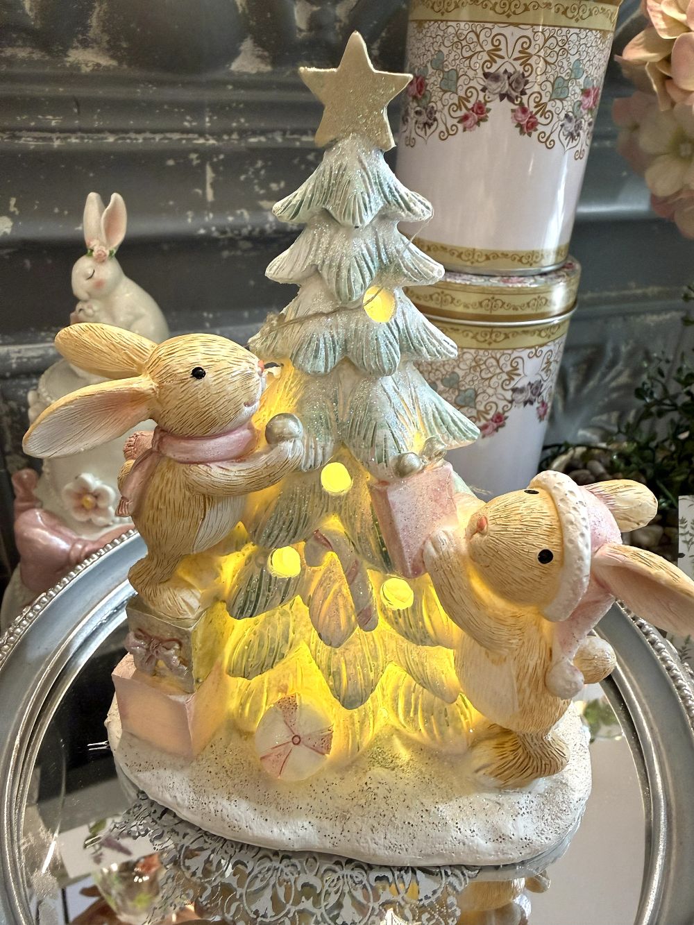 Figurine de Noël avec deux lapinous autour d’un sapin lumineux, tons vert givré, crème et rose poudré, décoration féérique et romantique