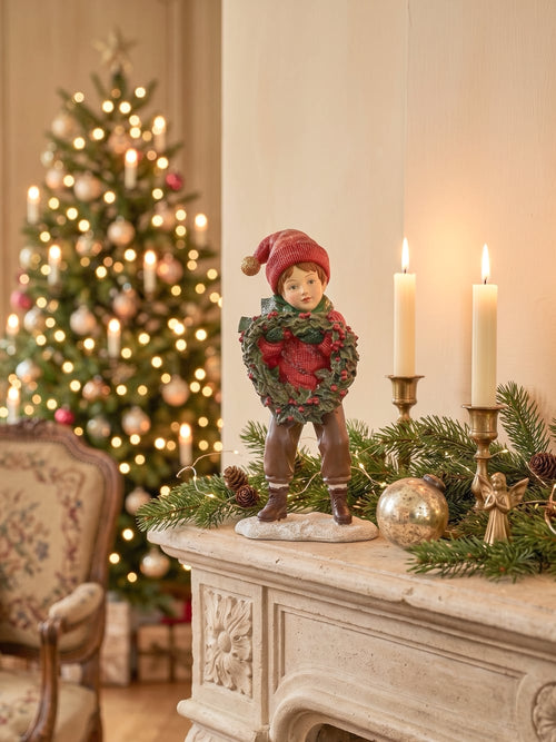 Figurine petit garçon de Noël en rouge et vert tenant une couronne végétale à baies rouges, décoration de Noël poétique et féérique