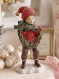 Figurine petit garçon de Noël en rouge et vert tenant une couronne végétale à baies rouges, décoration de Noël poétique et féérique