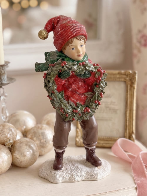 Figurine petit garçon de Noël en rouge et vert tenant une couronne végétale à baies rouges, décoration de Noël poétique et féérique