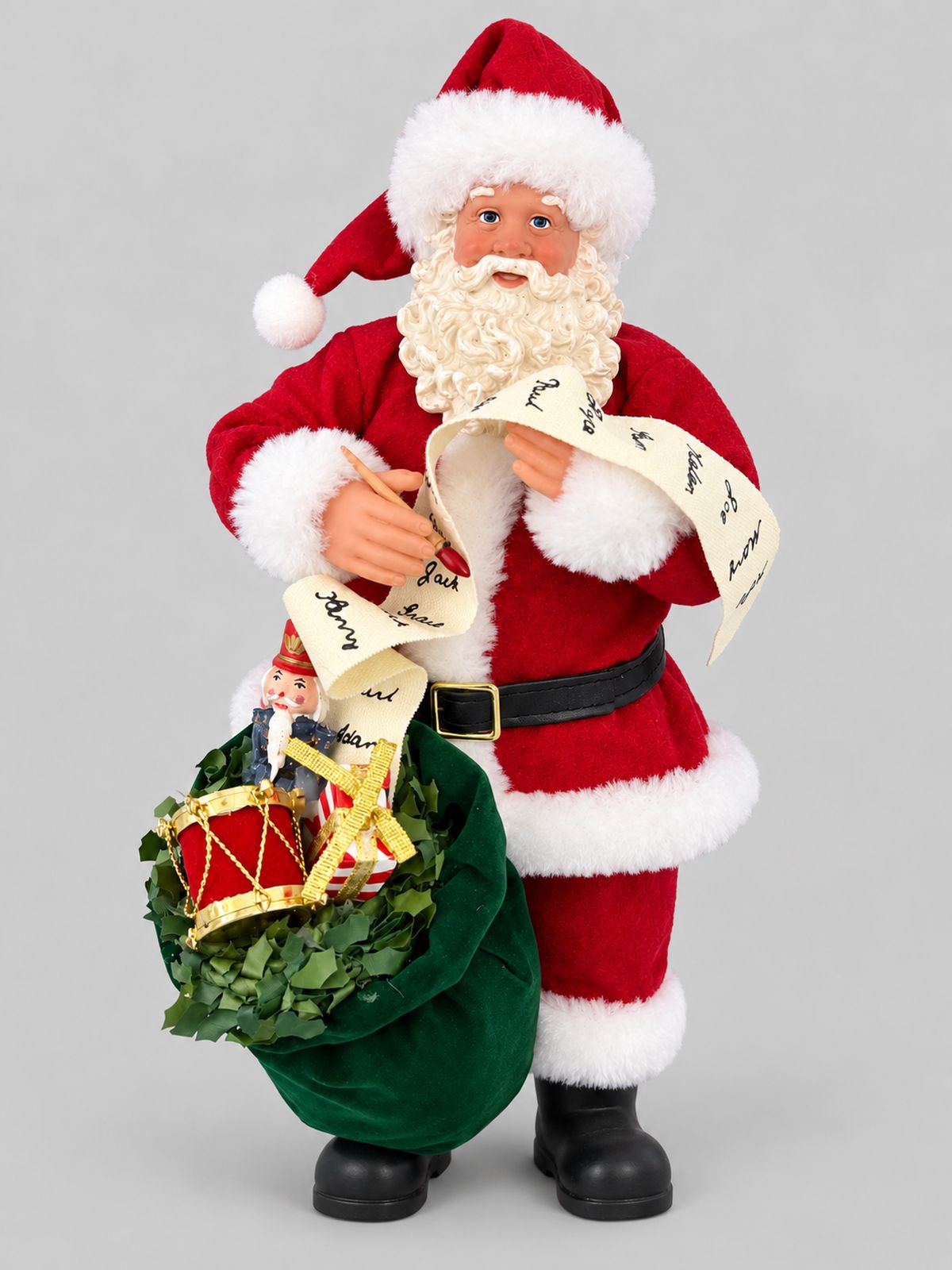 Figurine de Père Noël en costume rouge tenant une liste, accompagnée d’un sac vert rempli de jouets, sur fond gris clair.