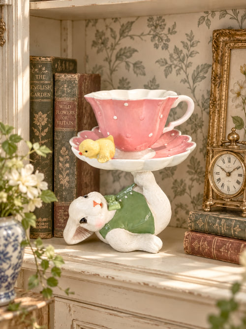 Figurine décorative avec lapin blanc vêtu de vert portant une grande tasse rose à pois, soucoupe fleurie et petit poussin jaune, ambiance de Pâques féérique