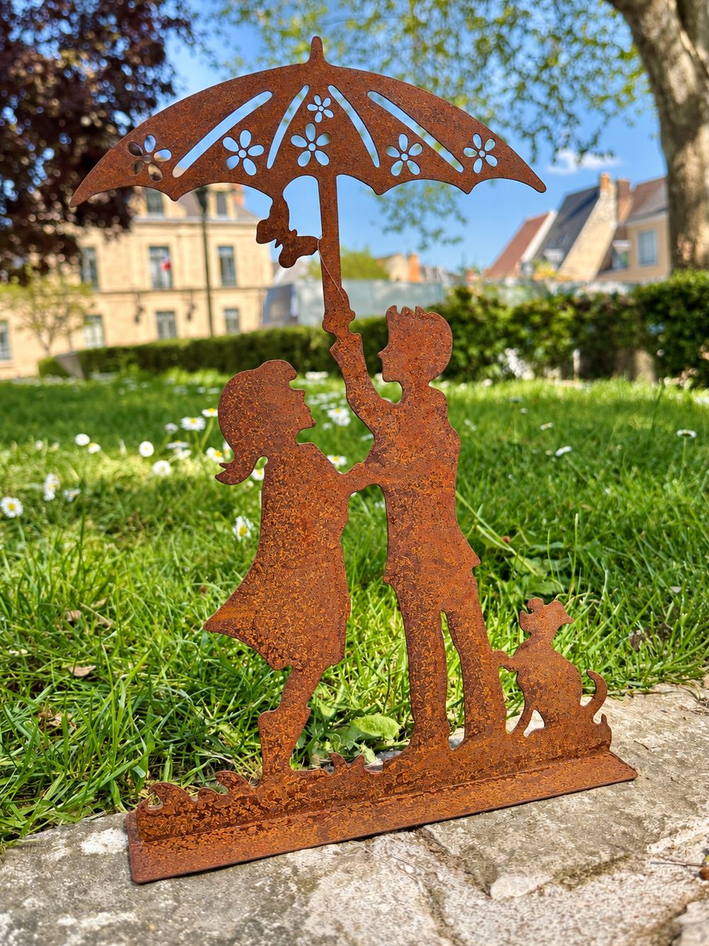 Silhouette Les Amoureux au Jardin sur un muret en pierre, devant une pelouse et des maisons anciennes.