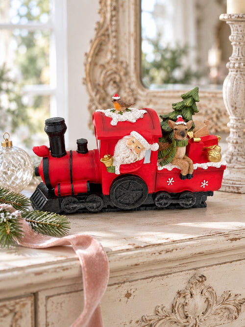 Locomotive de Noël rouge sur une console ivoire patinée, près d’une boule de verre et d’un ruban rose poudré.