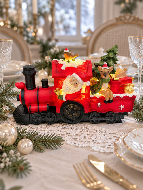 Locomotive de Noël allumée au centre d’une table de fête, entourée de porcelaine blanche et de branches de sapin.