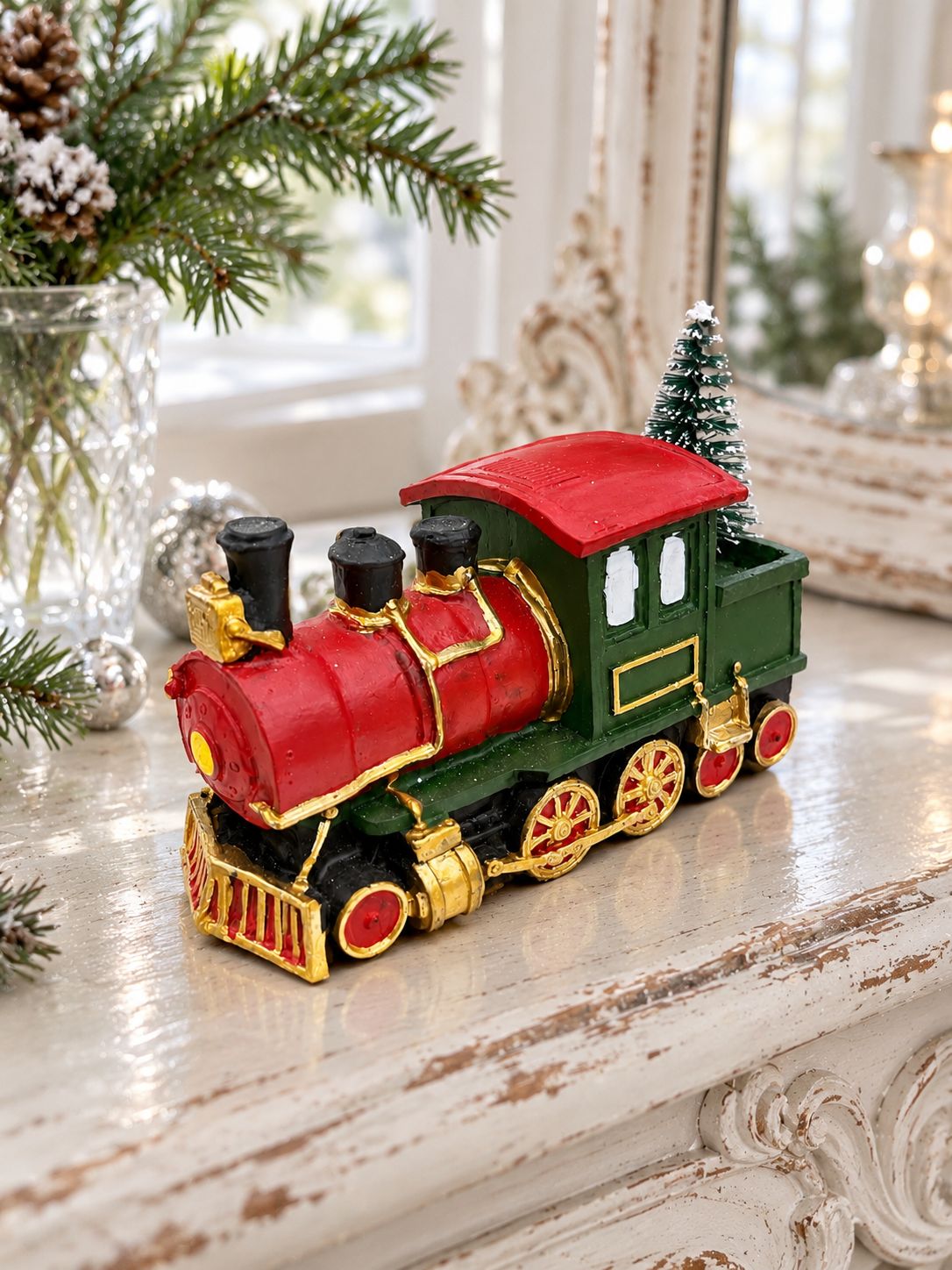 Locomotive miniature de Noël sur une console blanche patinée, près d’un miroir ancien et de branches de sapin.