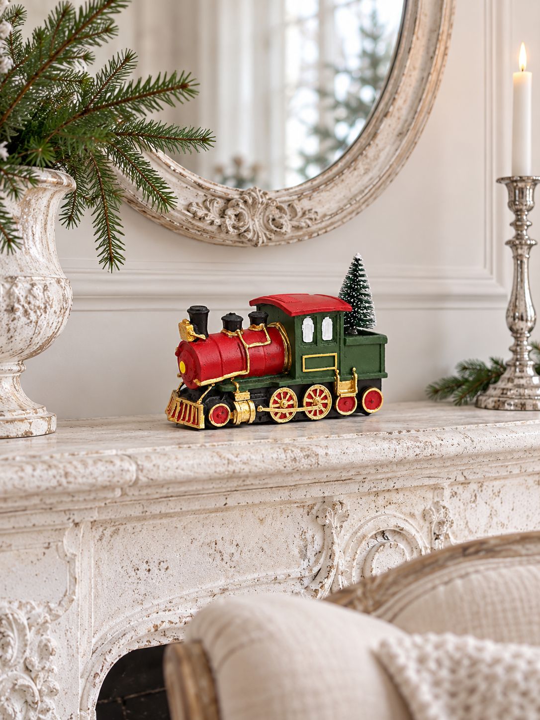 Petite locomotive de Noël sur une cheminée en pierre claire, sous un miroir ancien, entre branches de sapin et bougeoir argenté.