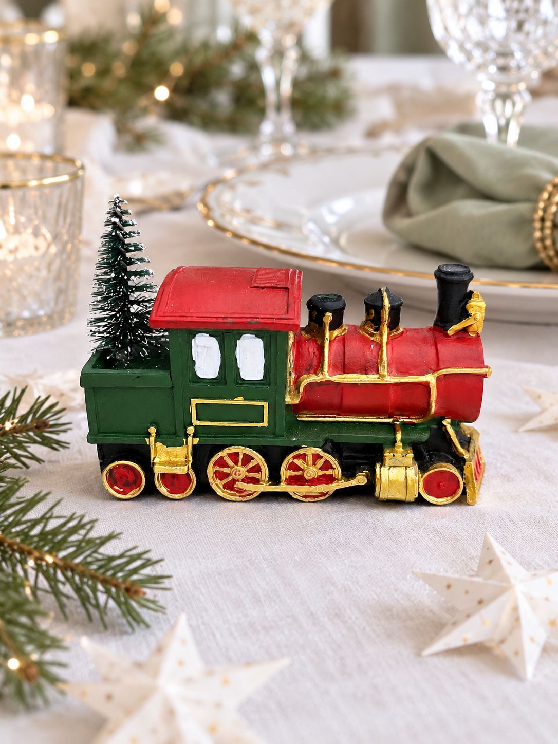 Locomotive décorative rouge et verte sur une table de Noël, avec nappe blanche, vaisselle dorée et étoiles en papier.