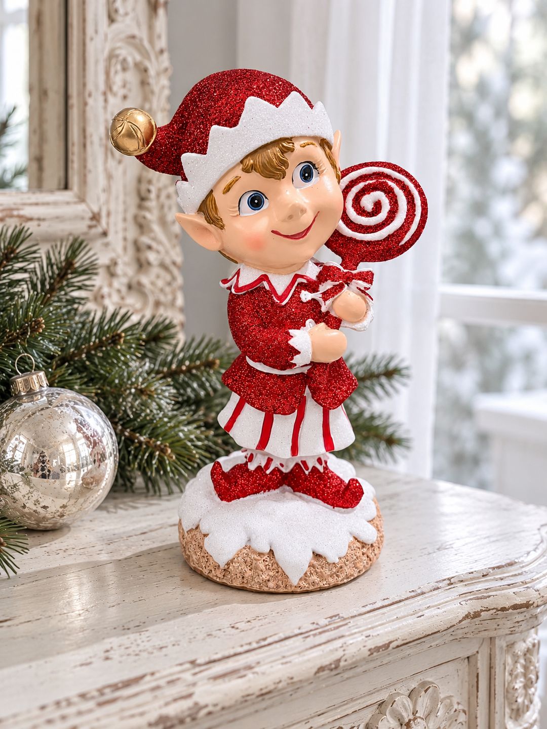 Lutin rouge pailleté sur une console blanche patinée près d’une fenêtre, accompagné de branches de sapin et d’une boule argentée.