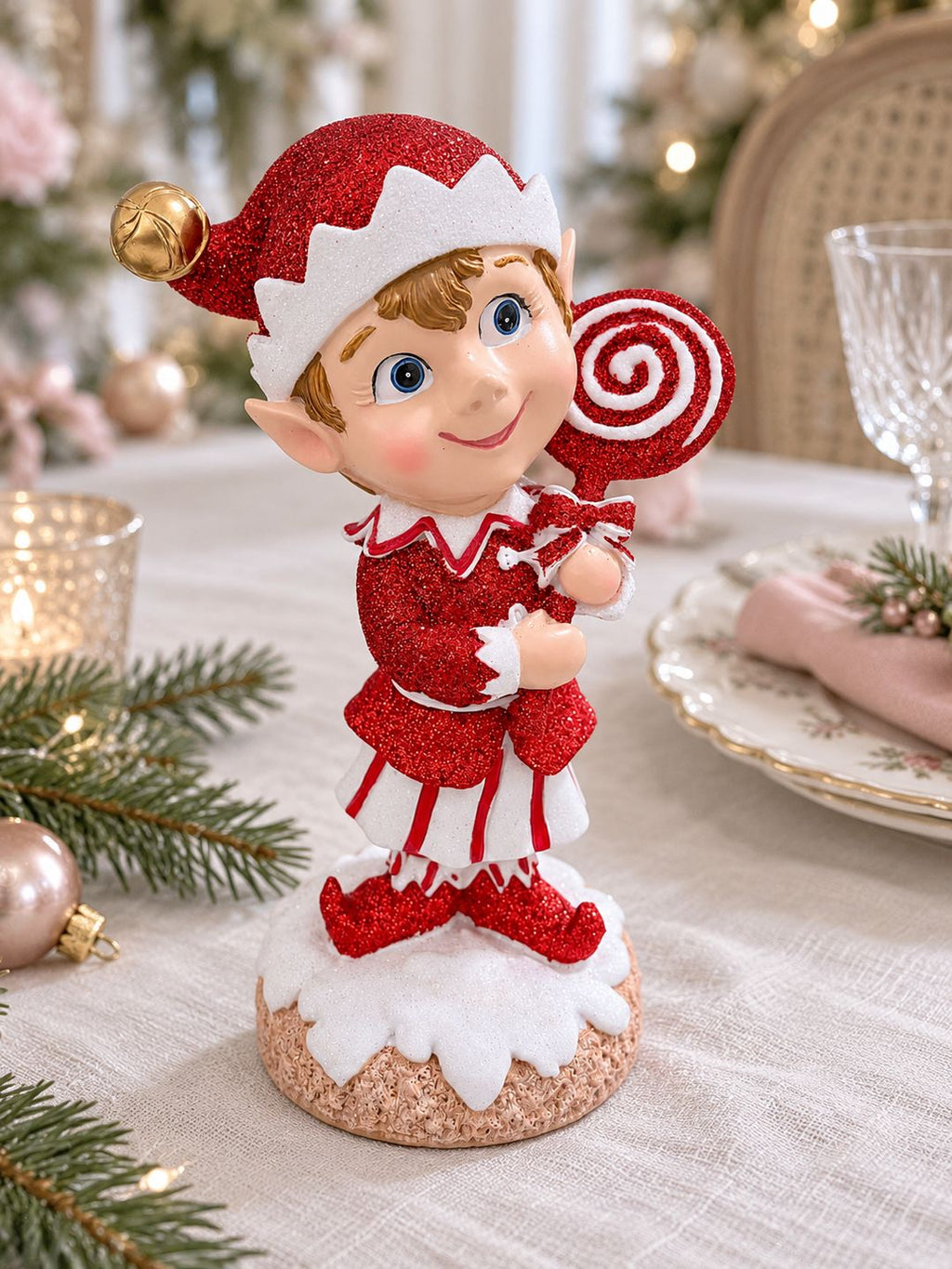 Lutin rouge pailleté au centre d’une table de Noël en lin clair, avec vaisselle fleurie et serviette rose poudré.