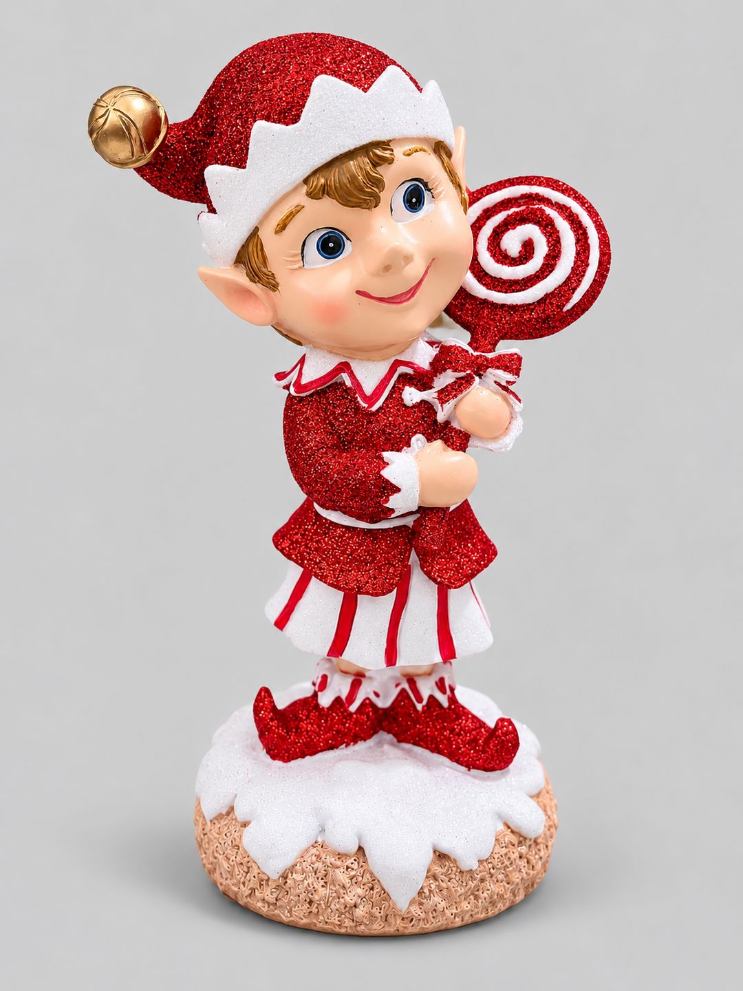 Figurine de lutin rouge pailleté tenant une sucette en spirale, sur un socle enneigé et un fond gris clair.
