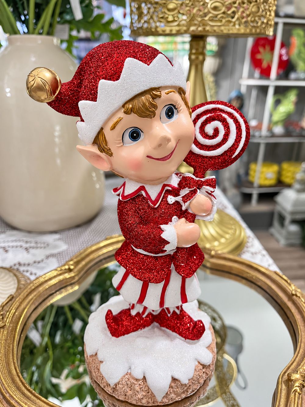 Lutin rouge pailleté vu de face, tenant une sucette rouge et blanche sur un socle enneigé.