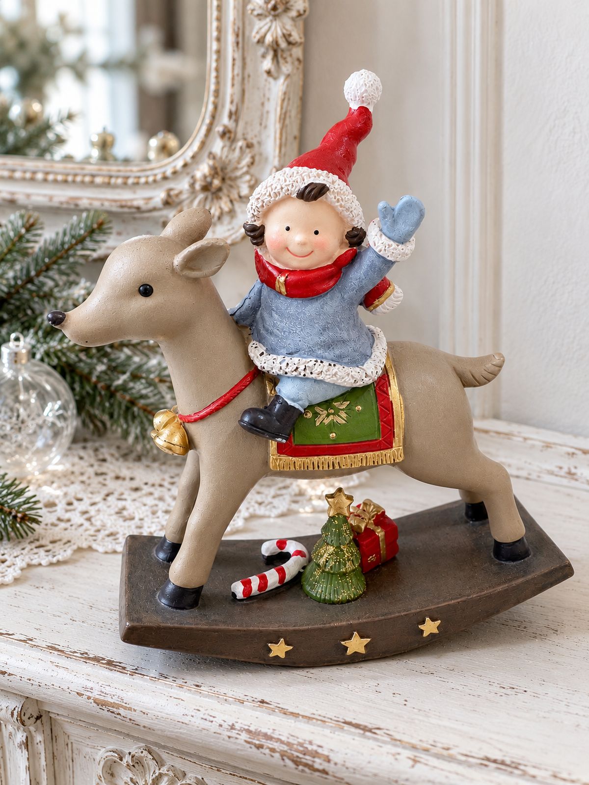 Figurine de lutin à dos de renne sur une console blanche patinée, devant un miroir ancien et quelques branches de sapin.