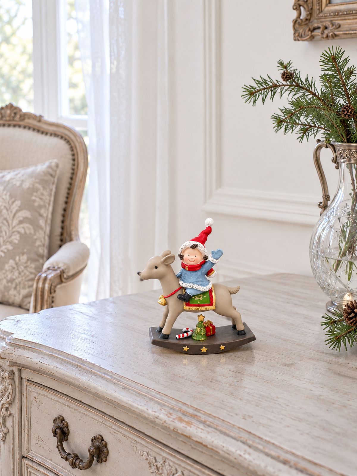 Petite figurine de lutin à dos de renne sur une commode patinée dans un intérieur lumineux, avec un fauteuil ancien et des branches de sapin.