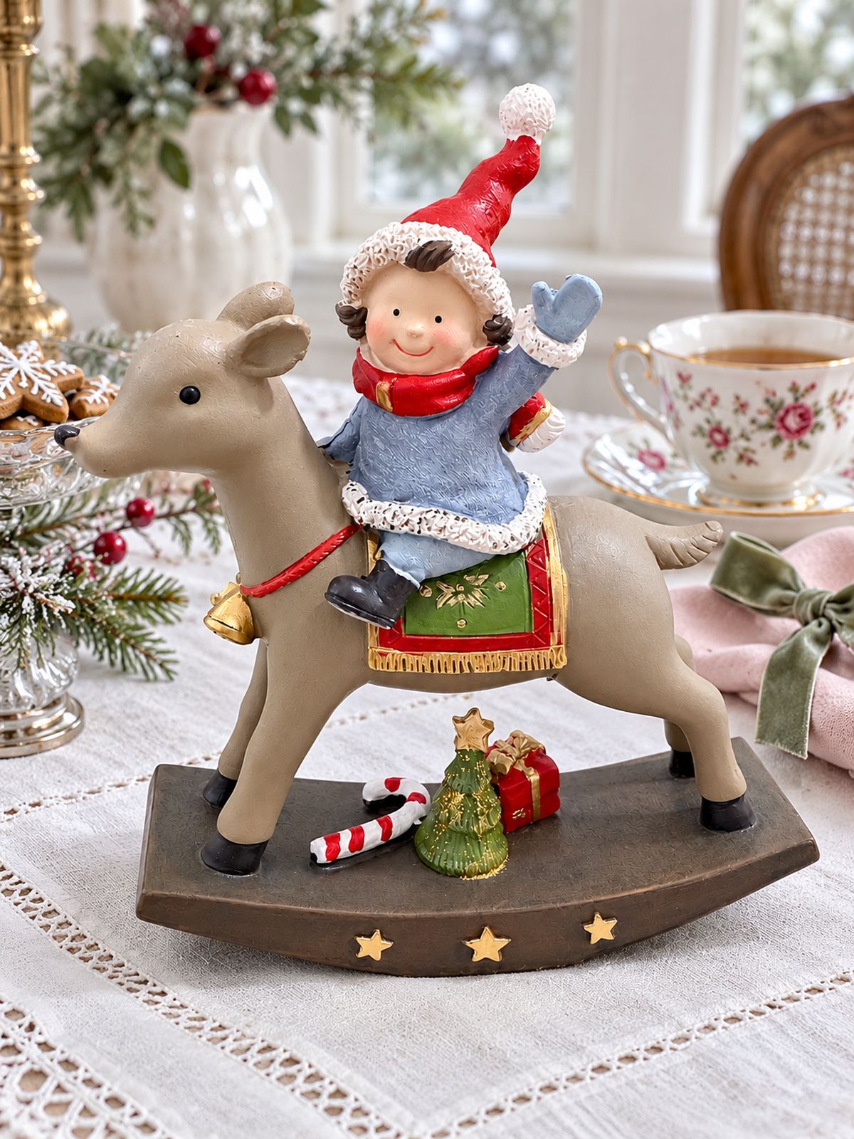 Figurine de lutin à dos de renne sur une table de Noël en lin blanc, près d’une tasse fleurie et d’une serviette rose.