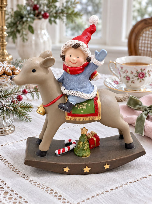 Figurine de lutin à dos de renne sur une table de Noël en lin blanc, près d’une tasse fleurie et d’une serviette rose.