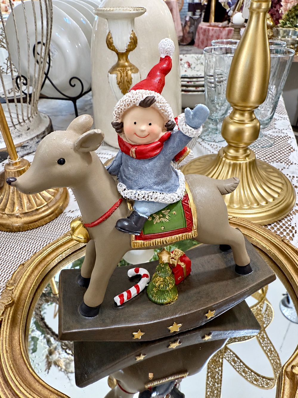 Figurine Lutin Joyeux à Dos de Renne vue de trois quarts, avec bonnet rouge, manteau bleu et socle décoré d’étoiles dorées.