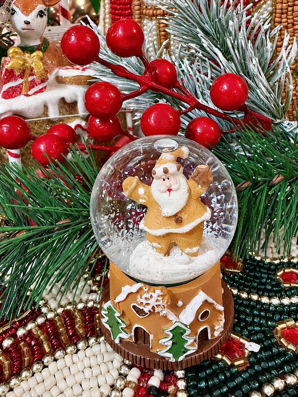 Mini boule à neige Père Noël Pain d’Épices, vue dans un décor de sapin et de baies rouges.