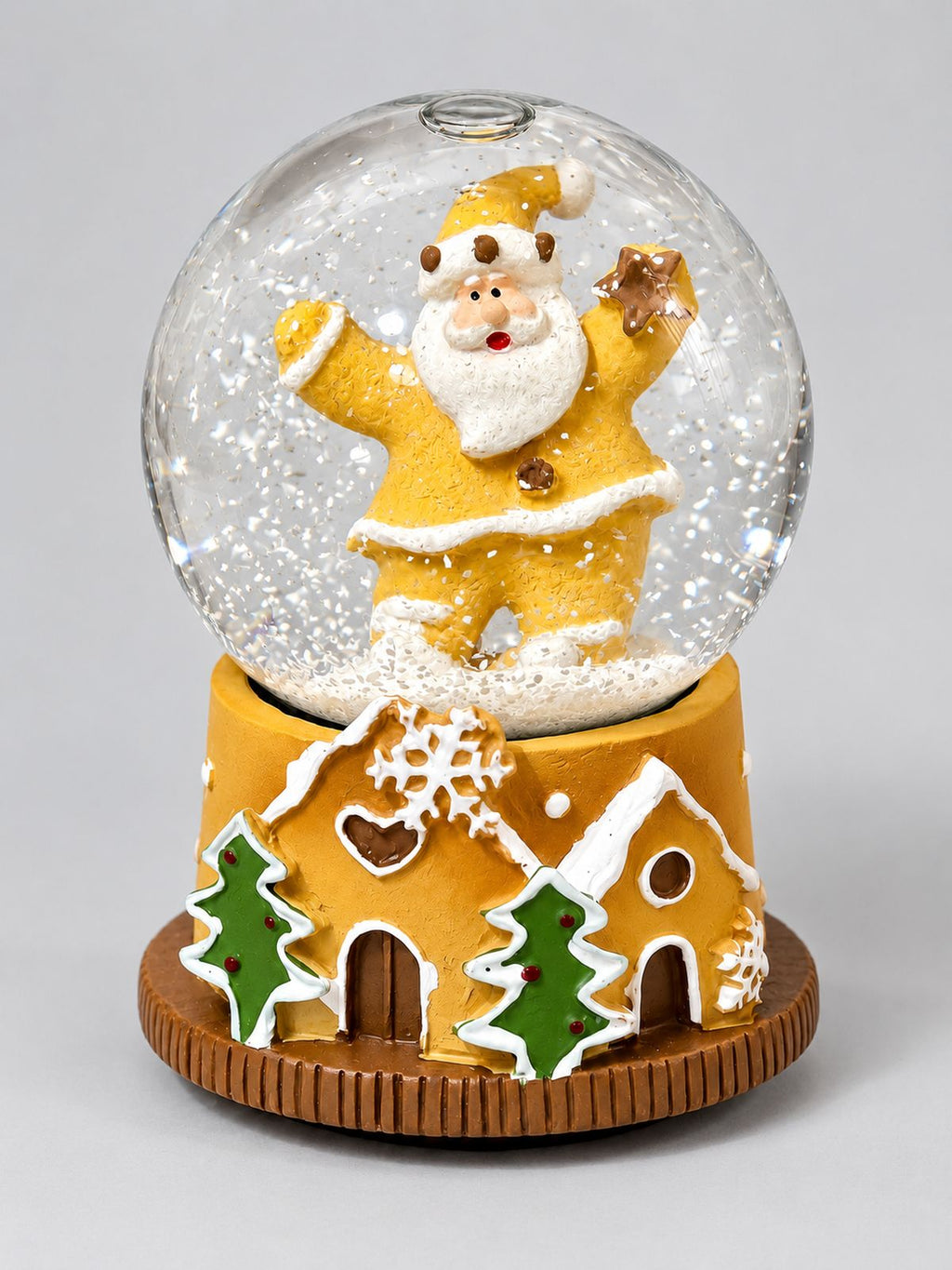 Mini boule à neige Père Noël Pain d’Épices, avec socle décoré de maisons et de sapins, sur fond gris clair.