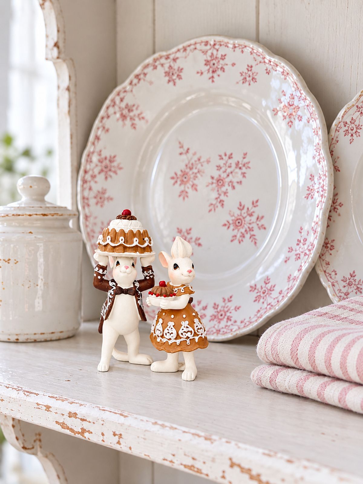 Duo Mr et Mme Pâtissouries sur une étagère patinée blanche, devant des assiettes anciennes à fleurs roses.