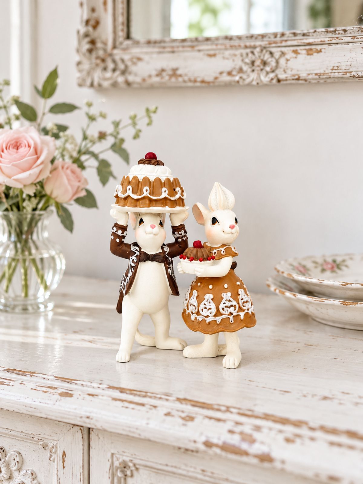 Figurines Mr et Mme Pâtissouries sur un buffet blanc patiné, entre un vase de roses et des assiettes anciennes.