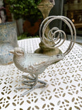 Oiseau décoratif en métal argent vieilli avec queue en spirale, style romantique et shabby chic, dimensions 18x10,5x20 cm, posé sur une table blanche ajourée.