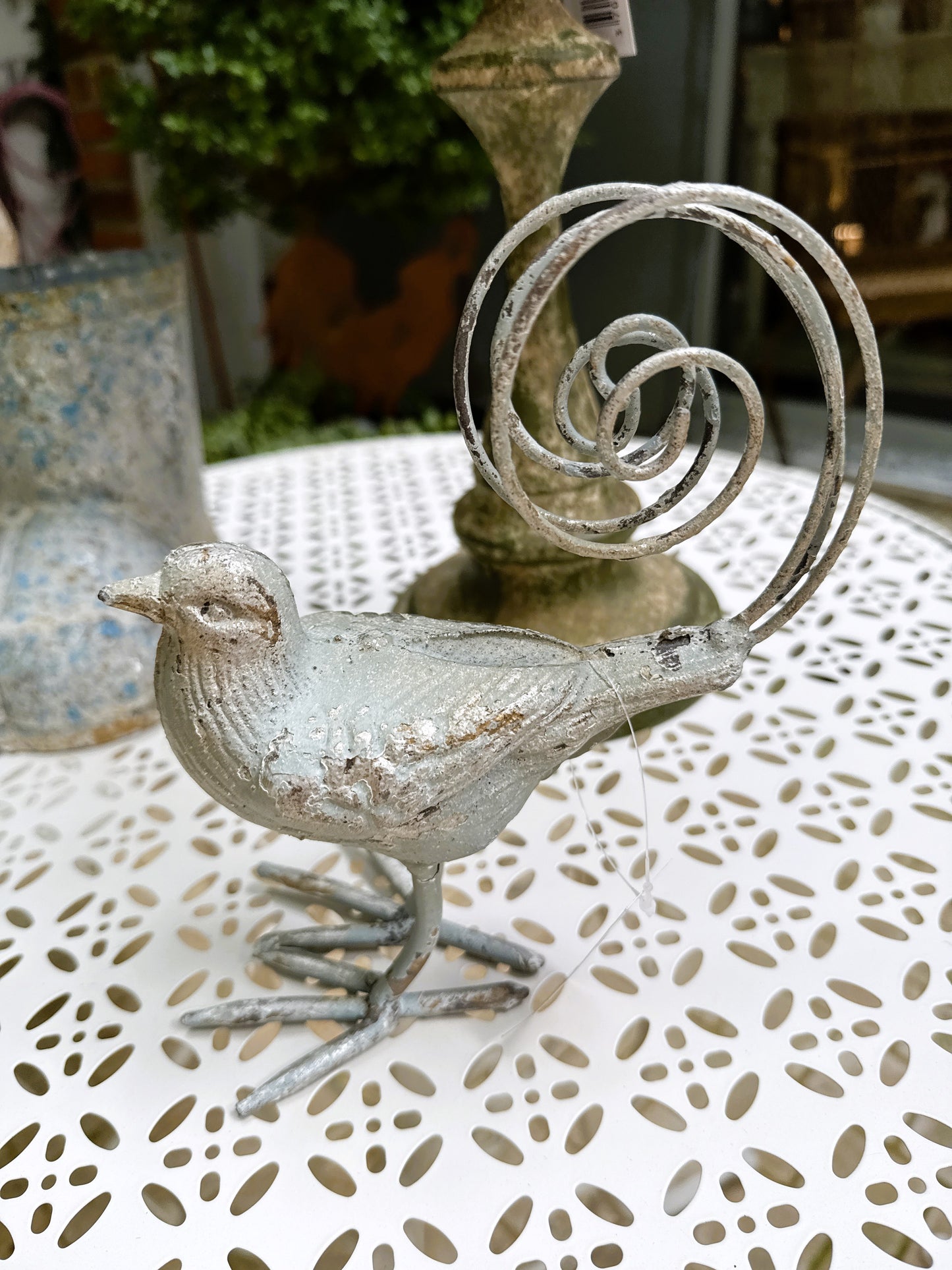 Oiseau décoratif en métal argent vieilli avec queue en spirale, style romantique et shabby chic, dimensions 18x10,5x20 cm, posé sur une table blanche ajourée.