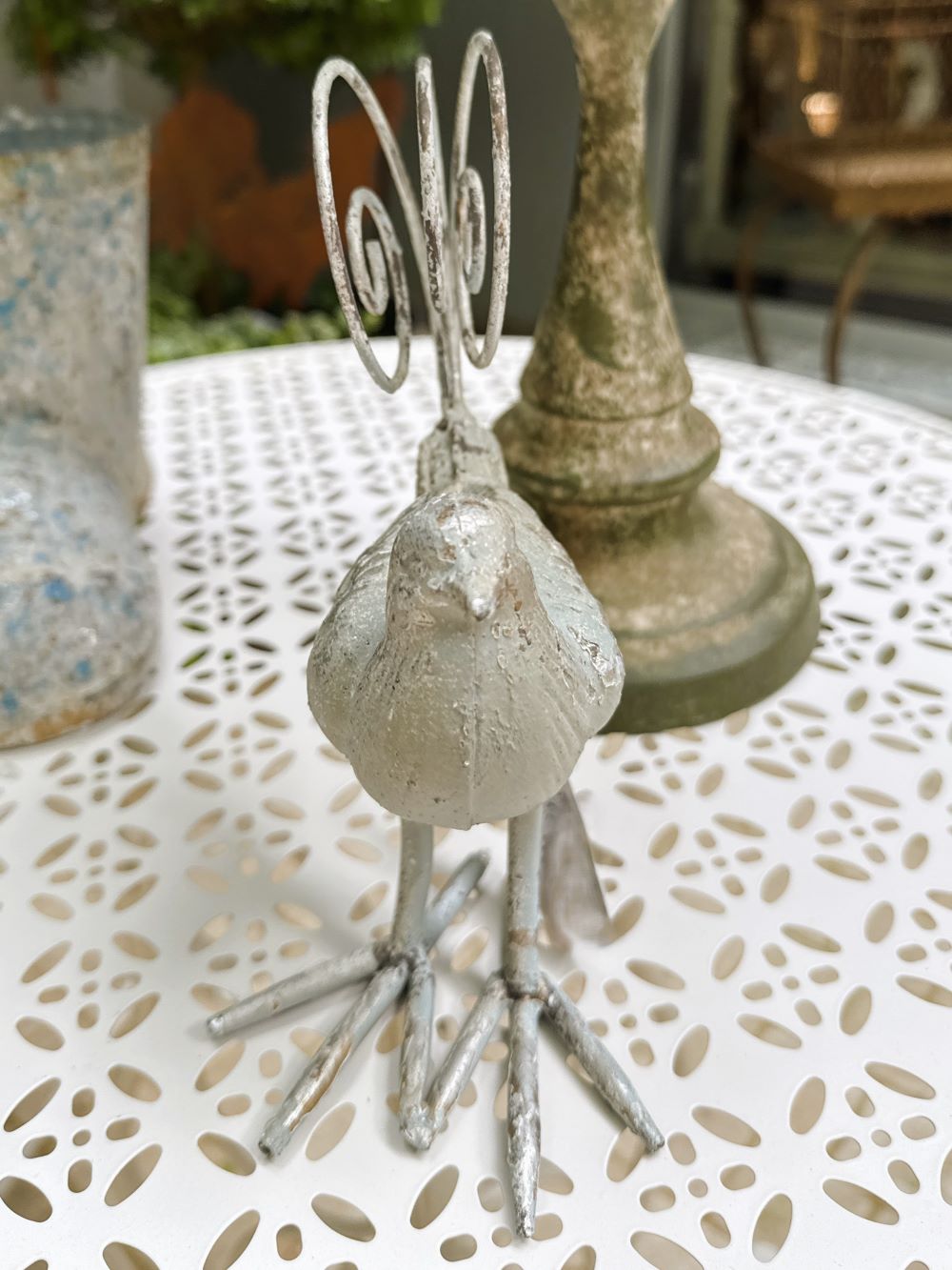 Oiseau décoratif en métal argent vieilli avec queue en spirale, style romantique et shabby chic, dimensions 18x10,5x20 cm, posé sur une table blanche ajourée.