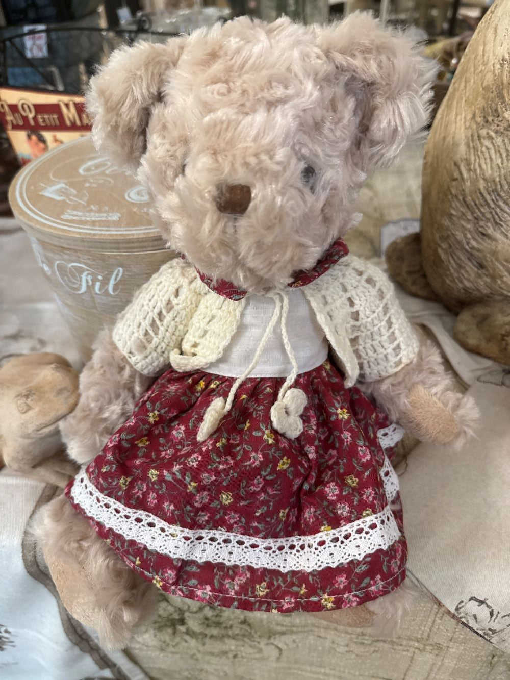 Oursonne décorative en peluche beige vêtue d’une robe fleurie rouge et d’un gilet crocheté écru, style shabby chic romantique pour chambre ou décoration de charme