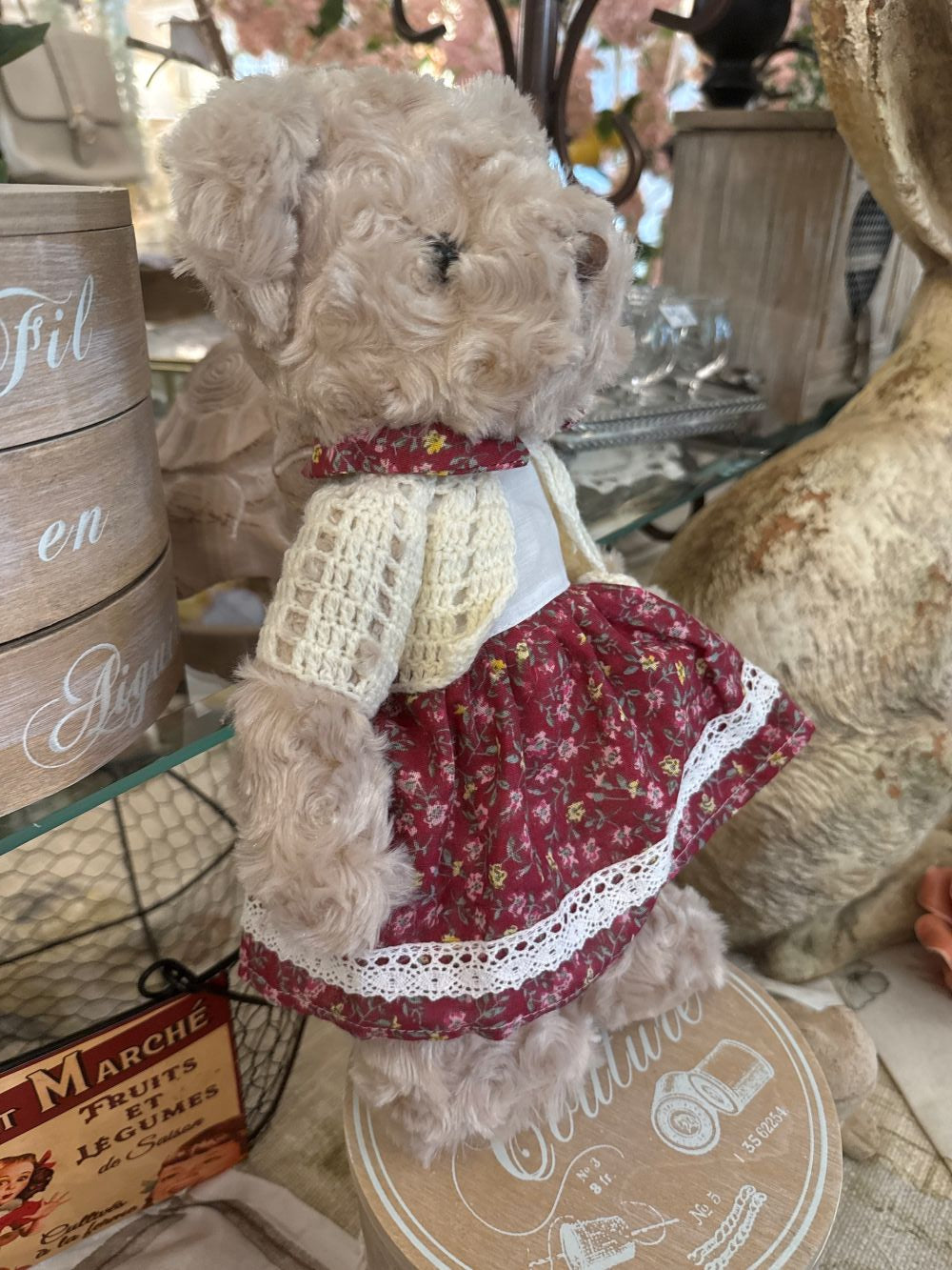 Oursonne décorative en peluche beige vêtue d’une robe fleurie rouge et d’un gilet crocheté écru, style shabby chic romantique pour chambre ou décoration de charme