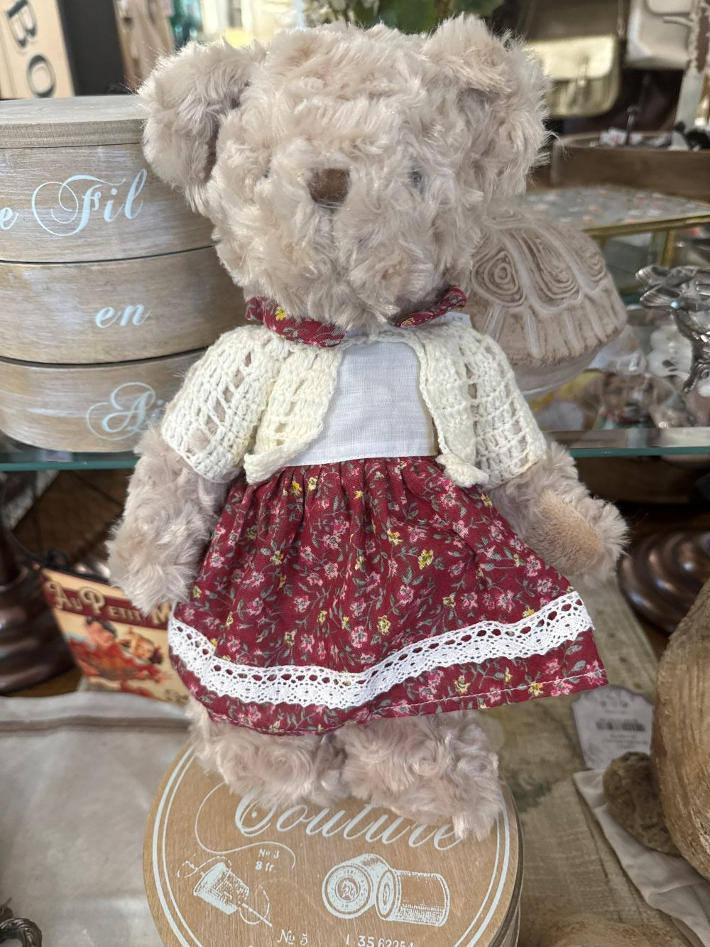 Oursonne décorative en peluche beige vêtue d’une robe fleurie rouge et d’un gilet crocheté écru, style shabby chic romantique pour chambre ou décoration de charme