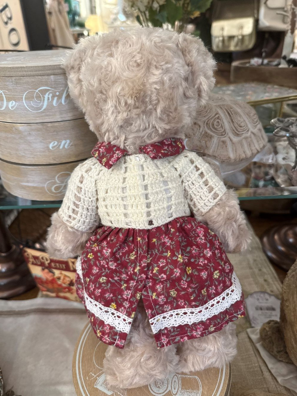 Oursonne décorative en peluche beige vêtue d’une robe fleurie rouge et d’un gilet crocheté écru, style shabby chic romantique pour chambre ou décoration de charme
