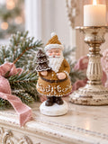 Père Noël Pain d’Épices Miniature sur une console ivoire patinée, près d’un bougeoir ancien, de branches de sapin et d’un ruban rose.