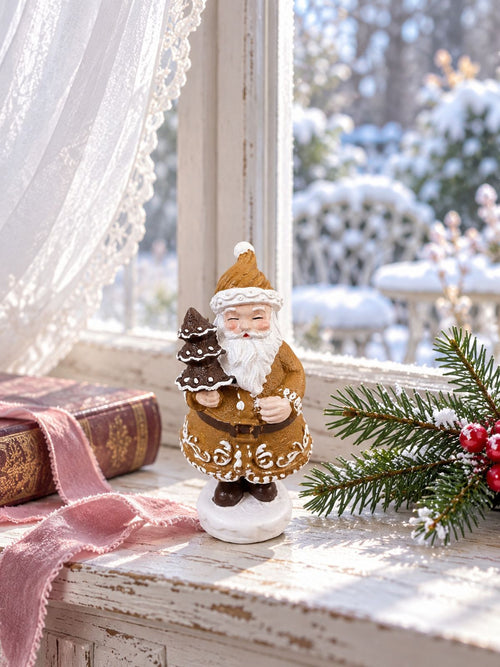 Père Noël Pain d’Épices Miniature devant une fenêtre donnant sur un jardin enneigé, accompagné d’un livre ancien et de branches de sapin.