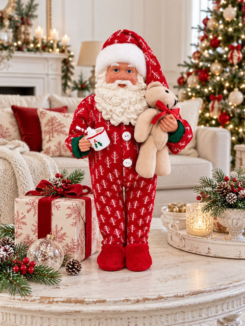 Père Noël en pyjama rouge avec son ourson, sur une table basse avec un cadeau, dans un salon décoré pour Noël.