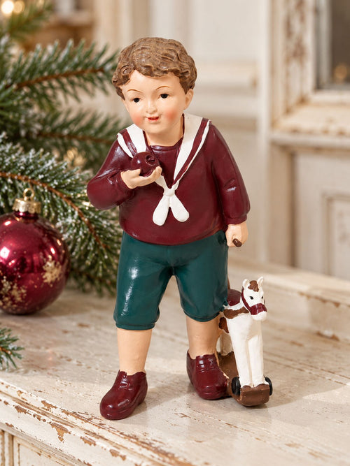 Figurine Petit Garçon au Cheval sur un meuble patiné, près de branches de sapin et d’une boule de Noël bordeaux.