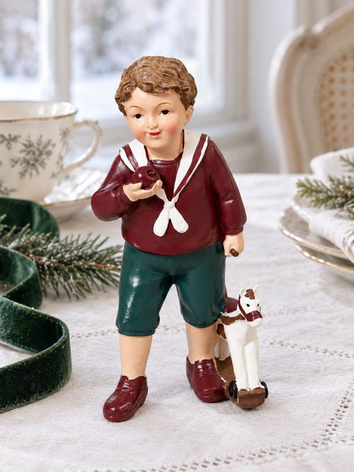 Figurine Petit Garçon au Cheval sur une table en lin blanc, avec une tasse ancienne et un ruban vert près d’une fenêtre.
