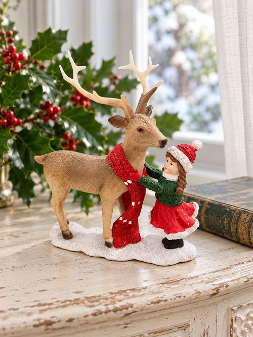 Figurine Petite Fille et Son Cerf sur une console patinée près d’une fenêtre, avec houx et livre ancien.