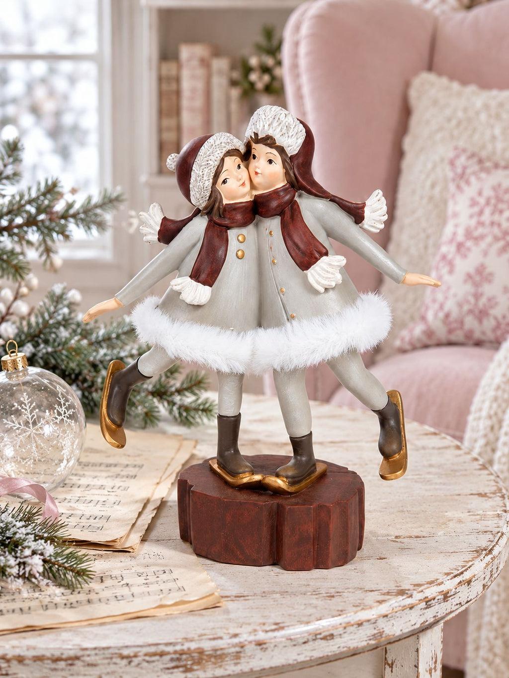 Figurine Les Petites Skieuses sur une table patinée avec des partitions anciennes et une boule de Noël en verre, devant un fauteuil rose poudré.