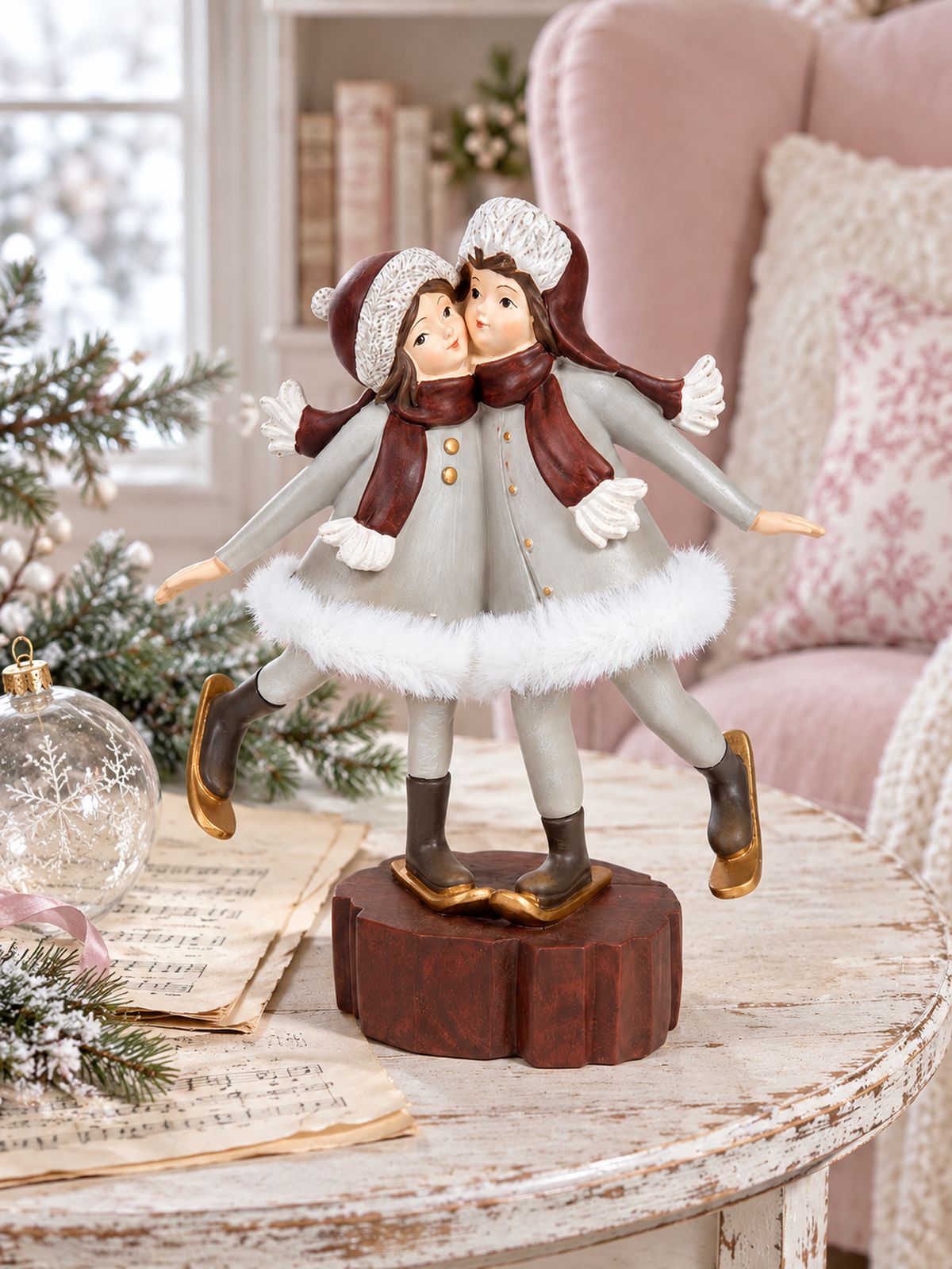 Figurine Les Petites Skieuses sur une table patinée avec des partitions anciennes et une boule de Noël en verre, devant un fauteuil rose poudré.