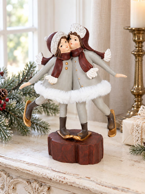 Figurine Les Petites Skieuses sur une console ivoire patinée près d’une fenêtre, accompagnée de branches de sapin et d’un bougeoir en laiton.