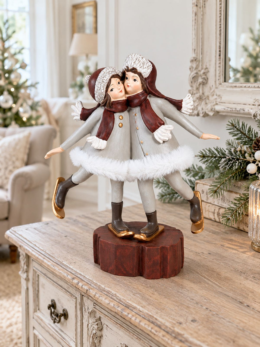 Figurine Les Petites Skieuses sur un buffet en bois patiné dans un salon lumineux, avec un miroir ancien et un sapin de Noël en arrière-plan.